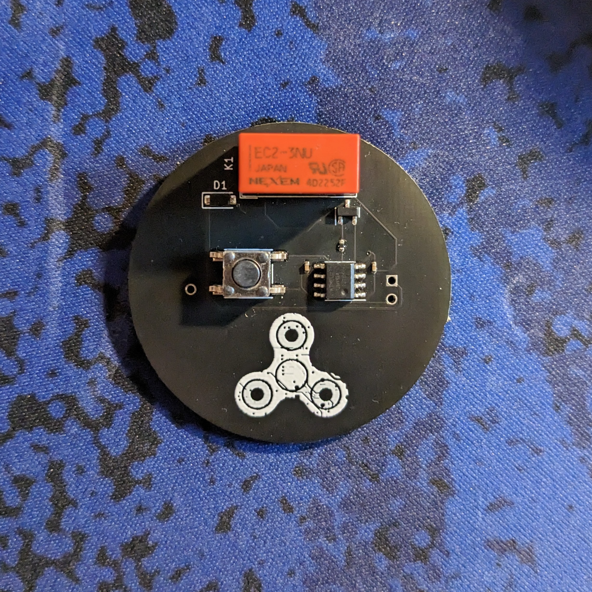 Version one of the E-Fidget Lite printed circuit board. Black board with an orange rectangular relay at the top and the E-Fidget logo in white silkscreen at the bottom.